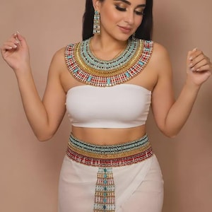 May include: A wide, multi-layered beaded collar necklace, matching drop earrings, and a coordinating wide beaded belt with a central hanging panel. The jewelry features intricate patterns of gold, turquoise, red, and white beads in a structured design.