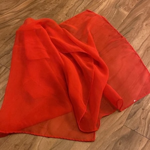 May include: A bright red sheer fabric scarf. The scarf is folded and draped on a wooden surface.