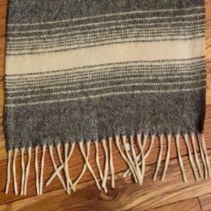 May include: A gray and cream-colored scarf with a herringbone pattern and a cream-colored stripe. The scarf has a fringe at the bottom. The scarf is made of a soft, woven material.