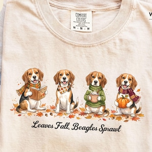 Może przedstawiać: Koszulka w kolorze kości słoniowej z grafiką przedstawiającą cztery psy rasy beagle siedzące na jesiennych liściach. Psy mają na sobie szaliki i trzymają przedmioty, w tym książkę, kubek i dynię. Tekst pod psami brzmi, Leaves Fall, Beagles Sprawl.
