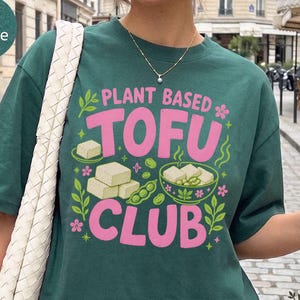Op de afbeelding: Een donkergroen t-shirt met de tekst "PLANT BASED TOFU CLUB" in grote roze letters. De afbeelding bevat illustraties van witte tofublokjes, groene sojabonen en kleine roze bloemetjes. Het t-shirt heeft een ronde hals en korte mouwen.