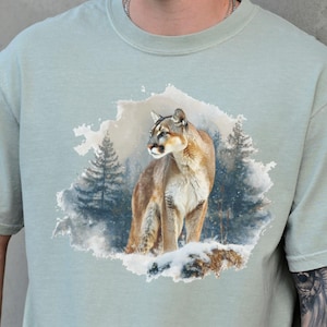 Puede incluir: Camiseta verde claro con una ilustración de un puma en una escena de bosque nevado. El puma está representado en tonos marrones y beige, con detalles de pelaje y rasgos faciales. La camiseta es de corte relajado.