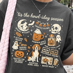 Könnte beinhalten: Ein anthrazitfarbenes T-Shirt mit dem Text 'tis the howl-iday season und verschiedenen Halloween-Illustrationen, darunter eine Hexe, ein Totenkopf, ein Kürbisgewürz-Becher, ein Beagle-Hund, Kassettendecks und Kürbisse in Orange, Weiß und Schwarz.