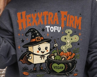 Puede incluir: Una sudadera gris jaspeado oscuro con cuello redondo que presenta un gráfico de un bloque de tofu con sombrero de bruja y capa, revolviendo un caldero negro con la etiqueta VEGAN y un corazón. El texto dice HEXXTRA FIRM TOFU en naranja, con estrellas.