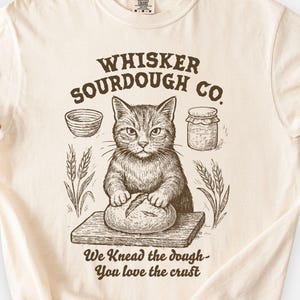 Op de afbeelding: Ivoor T-shirt met een kat die deeg kneedt, met de tekst "Whisker Sourdough Co." en "We Knead the dough - You love the crust." Het ontwerp bevat illustraties van een kom, pot en tarwe.
