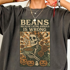 May include: Dark gray t-shirt with a graphic that reads "BEANS BECAUSE EATING ANIMALS IS WRONG." The graphic features a skeleton wearing a bandana, holding a bowl of beans, surrounded by pumpkins and roses. The shirt has short sleeves.