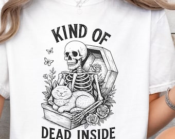 May include: A white t-shirt featuring a black graphic illustration of a skeleton sitting in a coffin with a cat. The text reads KIND OF DEAD INSIDE BUT A CAT PERSON. The design includes floral elements and butterflies surrounding the skeleton and cat.