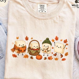 May include: An ivory t-shirt featuring a graphic of four smiling tofu blocks. One wears an orange scarf and holds a pumpkin, one sits in a woven basket with apples, one wears a green beanie and holds a mug, and one wears brown boots.