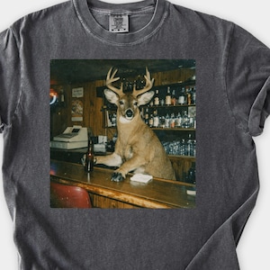 May include: Pepper-colored t-shirt featuring a square graphic of a deer standing behind a bar. The deer has antlers and is leaning on the bar. The bar has bottles and a cash register. The shirt has rolled sleeves and a crew neck. The Wild Paws Tees logo is on the bottom right.