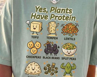 May include: A sage green t-shirt featuring the text Yes, Plants Have Protein and cartoon illustrations of tofu, tempeh, lentils, chickpeas, black beans, split peas, quinoa, hemp seeds, and pumpkin seeds with their names printed underneath each item.