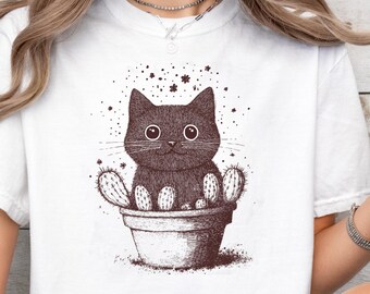 May include: White t-shirt featuring a brown illustration of a cat in a cactus pot. The cat has large eyes and whiskers. The design includes stars and small dots. The shirt is a casual style.