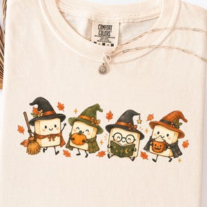 Può includere: Una t-shirt girocollo color avorio con una grafica raffigurante quattro blocchi di tofu in stile cartoon che indossano cappelli da strega. I personaggi sono ritratti con elementi autunnali come una scopa, una zucca, un libro e una tazza.