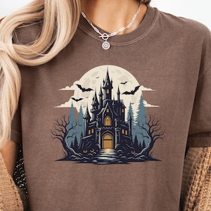 May include: Espresso-colored t-shirt featuring a spooky castle graphic. The design includes a full moon, bats, and trees, creating a Halloween theme. The shirt is a comfortable, casual wear item.