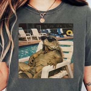May include: Dark gray t-shirt featuring a graphic of an iguana wearing sunglasses, lounging in a white chair by a pool. The iguana is the focal point, with a relaxed pose. The shirt is a casual, comfortable style.
