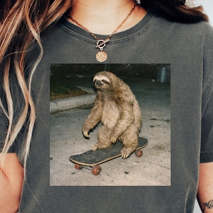 May include: Dark grey t-shirt featuring a square image of a sloth riding a skateboard. The sloth is brown and appears to be in motion. The skateboard is black with red wheels. The shirt is a casual style.