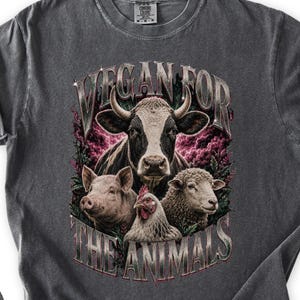May include: A charcoal gray short-sleeve t-shirt featuring a graphic print of a cow, pig, chicken, and sheep surrounded by pink clouds and foliage. The text VEGAN FOR THE ANIMALS is printed in a metallic-style font above and below the animals.