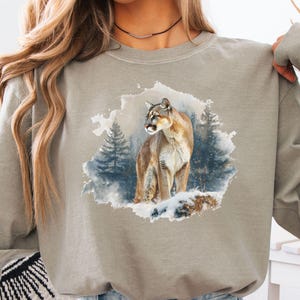 Puede incluir: Camiseta de manga larga color arenisca con una ilustración en acuarela de un puma en una escena de bosque nevado. El puma está representado en tonos marrones y beige, con un fondo de árboles azules y verdes. La camiseta es de material suave.