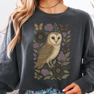 Puede incluir: Camiseta de manga larga gris oscuro con una ilustraci&oacute;n detallada de un b&uacute;ho com&uacute;n rodeado de flores y mariposas. El b&uacute;ho tiene un cuerpo marr&oacute;n claro y blanco con una cara en forma de coraz&oacute;n. Las flores son moradas y marr&oacute;n oscuro.