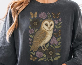 May include: Dark grey long-sleeve shirt featuring a detailed illustration of a barn owl surrounded by flowers and butterflies. The owl has a light brown and white body with a heart-shaped face. The flowers are purple and dark brown.