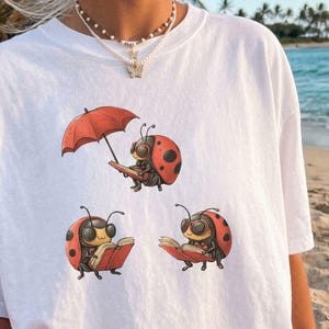 Op de afbeelding: Een wit t-shirt met een afbeelding van drie lieveheersbeestjes die boeken lezen, waarvan er één een rode paraplu vasthoudt. De tekst WILD PAWS TEES staat in de rechterbovenhoek. Het t-shirt heeft een ronde hals en korte mouwen.