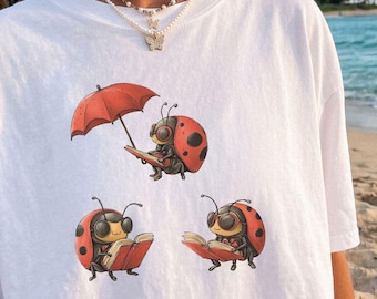 Könnte beinhalten: Ein weißes T-Shirt mit einer Grafik von drei Marienkäfern, die Bücher lesen, wobei ein Marienkäfer einen roten Regenschirm hält. Der Text WILD PAWS TEES ist in der oberen rechten Ecke gedruckt. Das T-Shirt hat einen Rundhalsausschnitt.