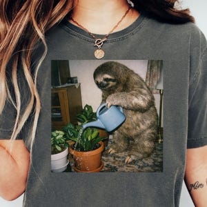 以下が含まれることがあります： 青いじょうろで植物に水をやっているナマケモノのグラフィックが特徴の濃い灰色のTシャツ。ナマケモノはキャビネットとランプのある部屋にいます。植物は茶色と白の鉢に入っています。