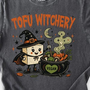 May include: A charcoal grey t-shirt featuring a graphic of a tofu block wearing a witch hat and cape, stirring a black cauldron labelled VEGAN with a heart. The text TOFU WITCHERY appears in orange above the illustration, which includes a crescent moon, stars, mushrooms, carrots, and broccoli.