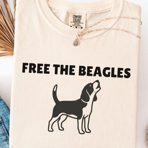 Może przedstawiać: Kremowa koszulka z napisem "FREE THE BEAGLES" i czarno-białą ilustracją beagle. Widoczny srebrny naszyjnik i niebieskie dżinsy. Koszulka jest złożona.