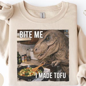 May include: Sand-colored sweatshirt with a humorous graphic of a Tyrannosaurus Rex cooking tofu. The text "BITE ME" is above the dinosaur, and "I MADE TOFU" is below. The sweatshirt has a crew neck and long sleeves.