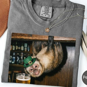 Può includere: T-shirt color pepe con una grafica di una scimmia con un cappello verde, appesa a testa in giù vicino a una birra. La maglietta ha un girocollo e maniche corte. Una collana d'argento è drappeggiata sulla maglietta.