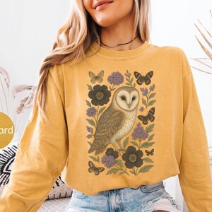 Puede incluir: Sudadera de color mostaza con una ilustraci&oacute;n detallada de un b&uacute;ho rodeado de flores y mariposas. El dise&ntilde;o incluye flores gris oscuro, acentos morados y mariposas negras. La palabra "Mustard" est&aacute; en un c&iacute;rculo en la parte inferior izquierda.