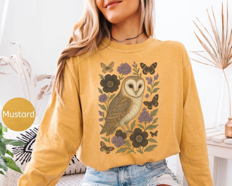 Puede incluir: Sudadera de color mostaza con una ilustraci&oacute;n detallada de un b&uacute;ho rodeado de flores y mariposas. El dise&ntilde;o incluye flores gris oscuro, acentos morados y mariposas negras. La palabra "Mustard" est&aacute; en un c&iacute;rculo en la parte inferior izquierda.