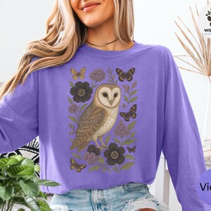 Puede incluir: Una sudadera violeta de manga larga con una ilustraci&oacute;n detallada de un b&uacute;ho rodeado de flores y mariposas. El dise&ntilde;o incluye el texto "Wild Paws Tees" y el nombre del color "Violet".