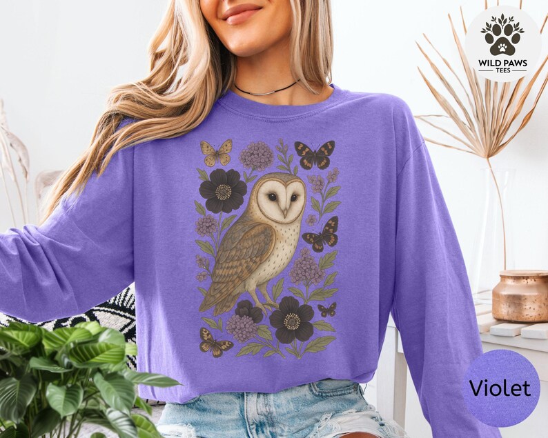 Puede incluir: Una sudadera violeta de manga larga con una ilustraci&oacute;n detallada de un b&uacute;ho rodeado de flores y mariposas. El dise&ntilde;o incluye el texto "Wild Paws Tees" y el nombre del color "Violet".