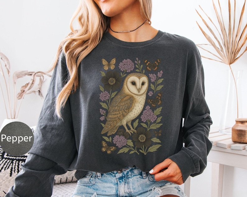 Puede incluir: Camiseta de manga larga gris oscuro con una ilustraci&oacute;n detallada de un b&uacute;ho com&uacute;n rodeado de flores y mariposas. El b&uacute;ho tiene un cuerpo marr&oacute;n claro y blanco con una cara en forma de coraz&oacute;n. Las flores son moradas y marr&oacute;n oscuro.