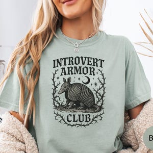 Op de afbeelding: Lichtgroen t-shirt met de tekst "INTROVERT ARMOR CLUB" die een gedetailleerde zwart-wit illustratie van een gordeldier omlijst. Het ontwerp bevat een maan en sterren.