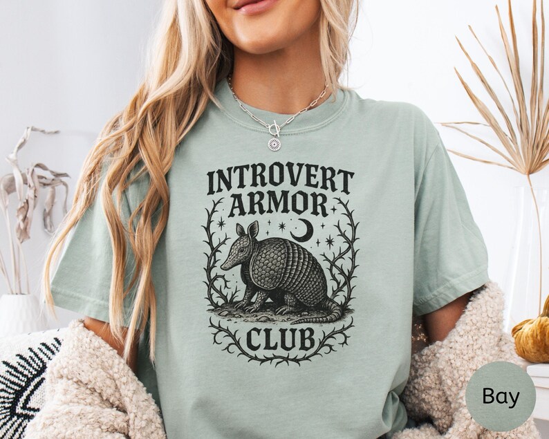 Op de afbeelding: Lichtgroen t-shirt met de tekst "INTROVERT ARMOR CLUB" die een gedetailleerde zwart-wit illustratie van een gordeldier omlijst. Het ontwerp bevat een maan en sterren.