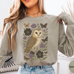 Puede incluir: Camiseta de manga larga color arena con una ilustraci&oacute;n detallada de un b&uacute;ho com&uacute;n rodeado de mariposas y flores en tonos marr&oacute;n, morado y negro. El b&uacute;ho es el punto focal, con intrincados detalles de plumas.