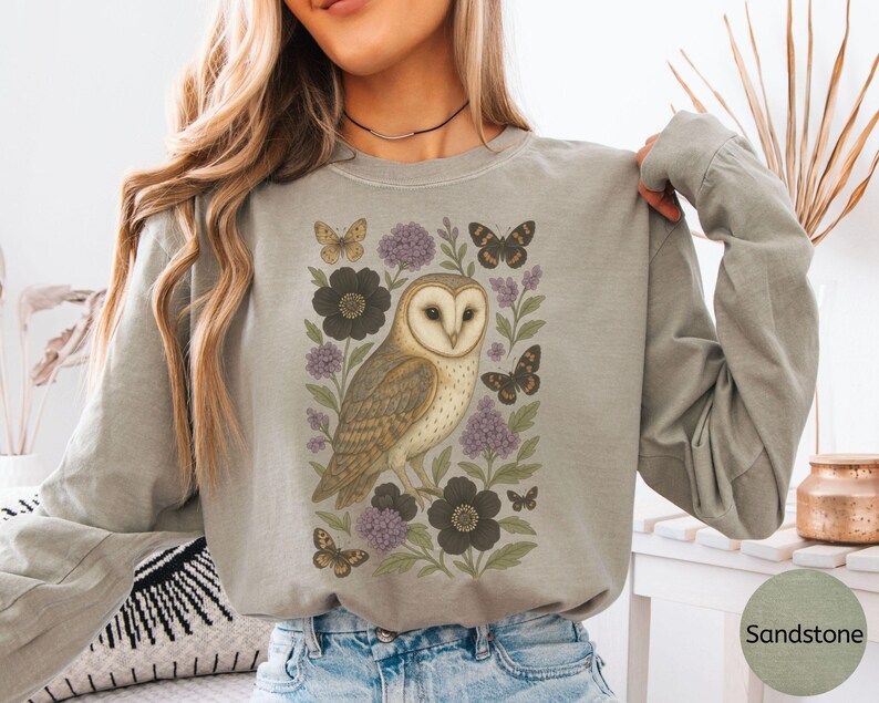 Puede incluir: Camiseta de manga larga color arena con una ilustraci&oacute;n detallada de un b&uacute;ho com&uacute;n rodeado de mariposas y flores en tonos marr&oacute;n, morado y negro. El b&uacute;ho es el punto focal, con intrincados detalles de plumas.