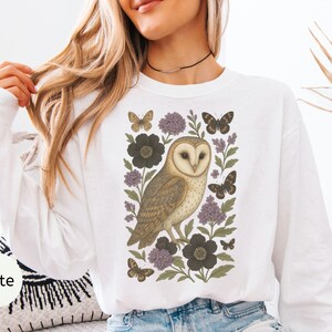 Puede incluir: Sudadera blanca de manga larga con una ilustraci&oacute;n detallada de un b&uacute;ho com&uacute;n rodeado de flores y mariposas en tonos marrones, negros y morados. La palabra "White" est&aacute; impresa en la parte inferior izquierda.