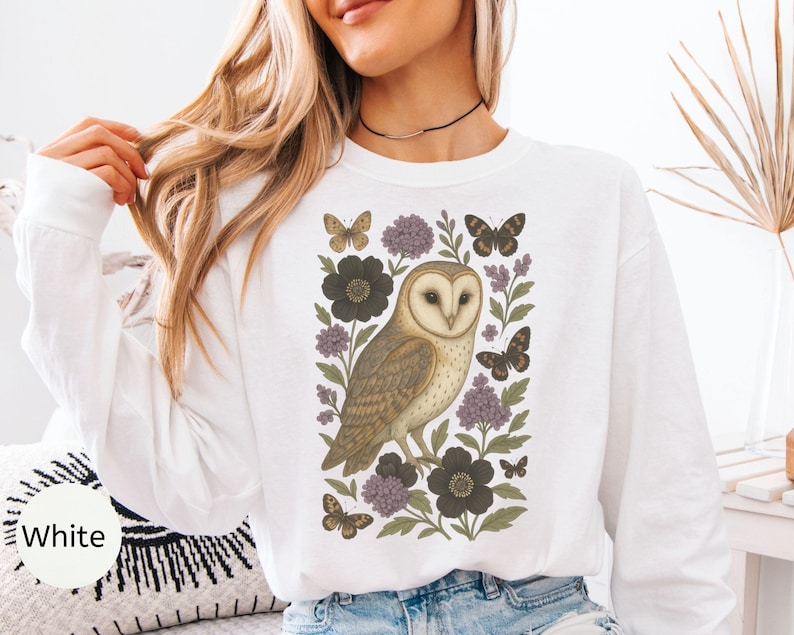 Puede incluir: Sudadera blanca de manga larga con una ilustraci&oacute;n detallada de un b&uacute;ho com&uacute;n rodeado de flores y mariposas en tonos marrones, negros y morados. La palabra "White" est&aacute; impresa en la parte inferior izquierda.