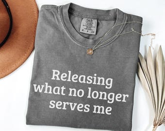 May include: A grey Comfort Colors t-shirt with the text "Releasing what no longer serves me" in white. A gold pendant necklace and a brown hat are also in the image. The shirt is folded and has a tag.