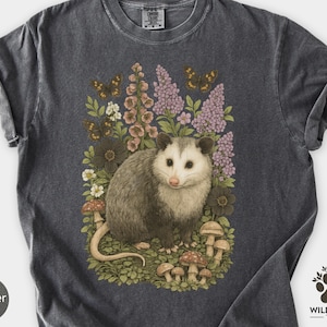 Puede incluir: Camiseta gris oscuro con una ilustración detallada de una zarigüeya rodeada de flores, mariposas y setas. La zarigüeya tiene pelaje gris y una nariz rosada. La camiseta tiene las mangas enrolladas y la palabra "Pepper" está impresa abajo a la izquierda.