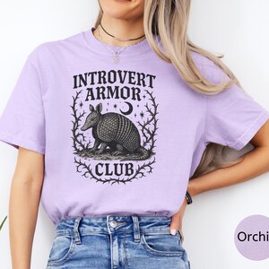 Op de afbeelding: Orchidee-kleurig T-shirt met de tekst "Introvert Armor Club" en een gedetailleerde illustratie van een gordeldier in een decoratief kader. Het ontwerp is in zwart en wit, wat een opvallend contrast cre&euml;ert met de zachte kleur van het shirt.