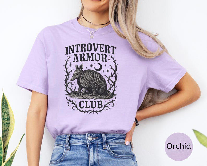 Op de afbeelding: Orchidee-kleurig T-shirt met de tekst "Introvert Armor Club" en een gedetailleerde illustratie van een gordeldier in een decoratief kader. Het ontwerp is in zwart en wit, wat een opvallend contrast cre&euml;ert met de zachte kleur van het shirt.