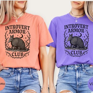 Op de afbeelding: Twee T-shirts, &eacute;&eacute;n terracotta en &eacute;&eacute;n violet, met de tekst "Introvert Armor Club" en een afbeelding van een gordeldier. Het gordeldier is omcirkeld door een krans van doornige wijnranken, een halve maan en sterren. Beide shirts worden gedragen met blauwe spijkerbroekshorts.