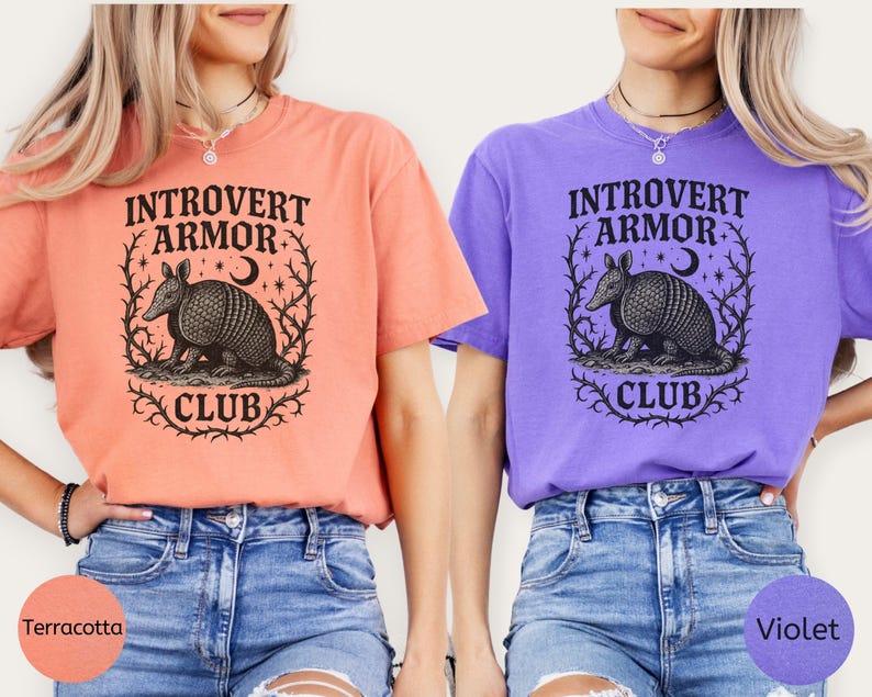Op de afbeelding: Twee T-shirts, &eacute;&eacute;n terracotta en &eacute;&eacute;n violet, met de tekst "Introvert Armor Club" en een afbeelding van een gordeldier. Het gordeldier is omcirkeld door een krans van doornige wijnranken, een halve maan en sterren. Beide shirts worden gedragen met blauwe spijkerbroekshorts.