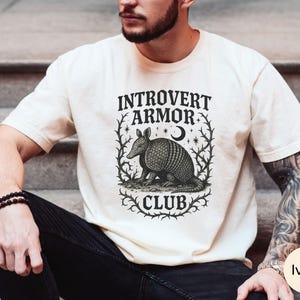 Op de afbeelding: Ivoor kleurig t-shirt met de tekst "Introvert Armor Club" en een gedetailleerde illustratie van een gordeldier. Het ontwerp is omlijst door een decoratieve rand met sterren en een maan.