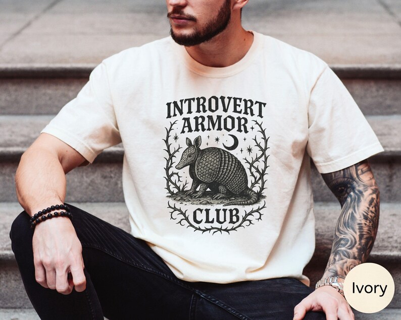 Op de afbeelding: Ivoor kleurig t-shirt met de tekst "Introvert Armor Club" en een gedetailleerde illustratie van een gordeldier. Het ontwerp is omlijst door een decoratieve rand met sterren en een maan.