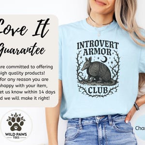 Op de afbeelding: Lichtblauw T-shirt met de tekst "INTROVERT ARMOR CLUB" en een gedetailleerde illustratie van een gordeldier. Het shirt heeft ook het "WILD PAWS TEES" logo en het woord "Chambray".